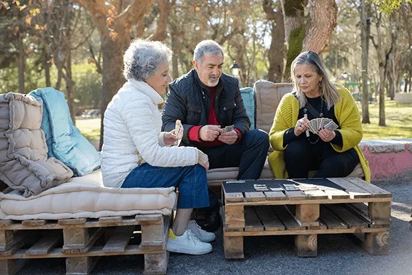 Three older adults sit on cushioned wooden pallet benches outdoors at MorningStar Senior Living Hillsboro, playing cards together on a low table. Trees and park surroundings offer a tranquil retirement living atmosphere in the background.