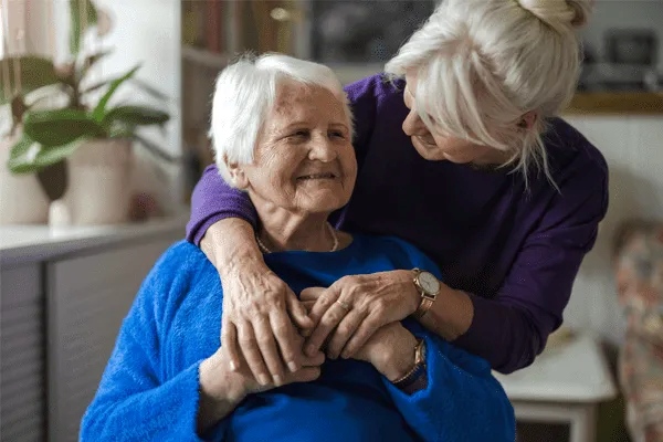 An elderly woman in a blue sweater smiles as a younger woman with gray hair embraces her from behind, both sharing a warm, affectionate moment at MorningStar Senior Living Hillsboro.