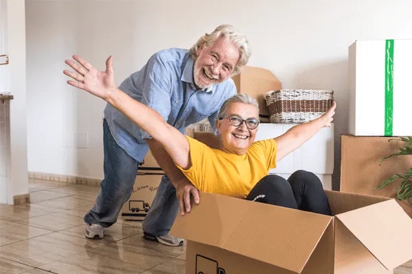 An older man smiles and stands behind an older woman sitting joyfully inside a cardboard box with her arms outstretched; moving boxes and baskets fill the background, capturing the excitement of starting life at MorningStar Senior Living Hillsboro.