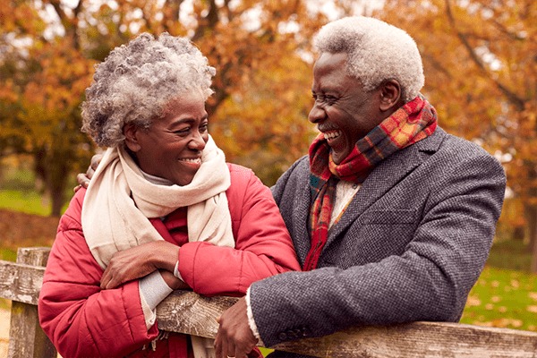 An older couple wearing warm coats and scarves smiles at each other while leaning on a wooden fence in a park with autumn leaves, capturing the warmth and connection found at MorningStar Senior Living Hillsboro's Retirement Living community.