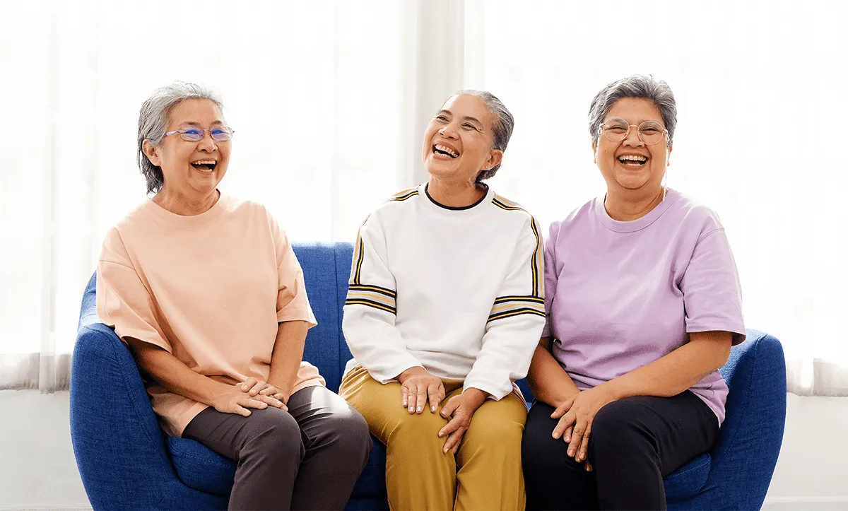Three older women sit on a blue couch and laugh together, dressed casually in pastel shirts and pants, enjoying each other's company at MorningStar Senior Living Hillsboro with a bright window in the background.