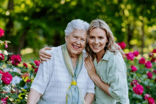 Two women, one elderly and one younger, smile and embrace in a garden filled with blooming pink and red flowers at MorningStar Senior Living Hillsboro, surrounded by green trees—a perfect snapshot of joyful retirement living.