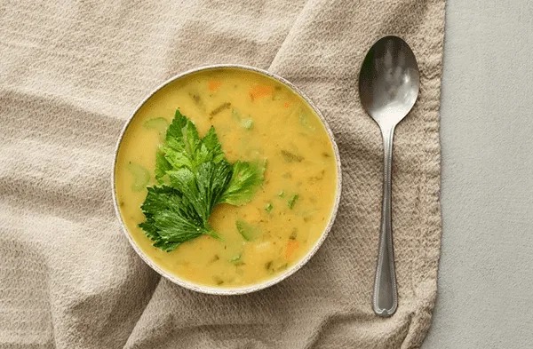 A bowl of creamy yellow vegetable soup garnished with fresh celery leaves, placed on a textured beige cloth next to a silver spoon, evokes the warm hospitality found at MorningStar Senior Living Hillsboro’s retirement living community.
