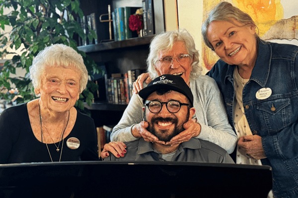 Three elderly women from MorningStar Senior Living Hillsboro smile behind a younger man seated at a piano. One playfully holds his face, as they all enjoy a happy moment in a cozy, warmly lit room filled with books and plants.