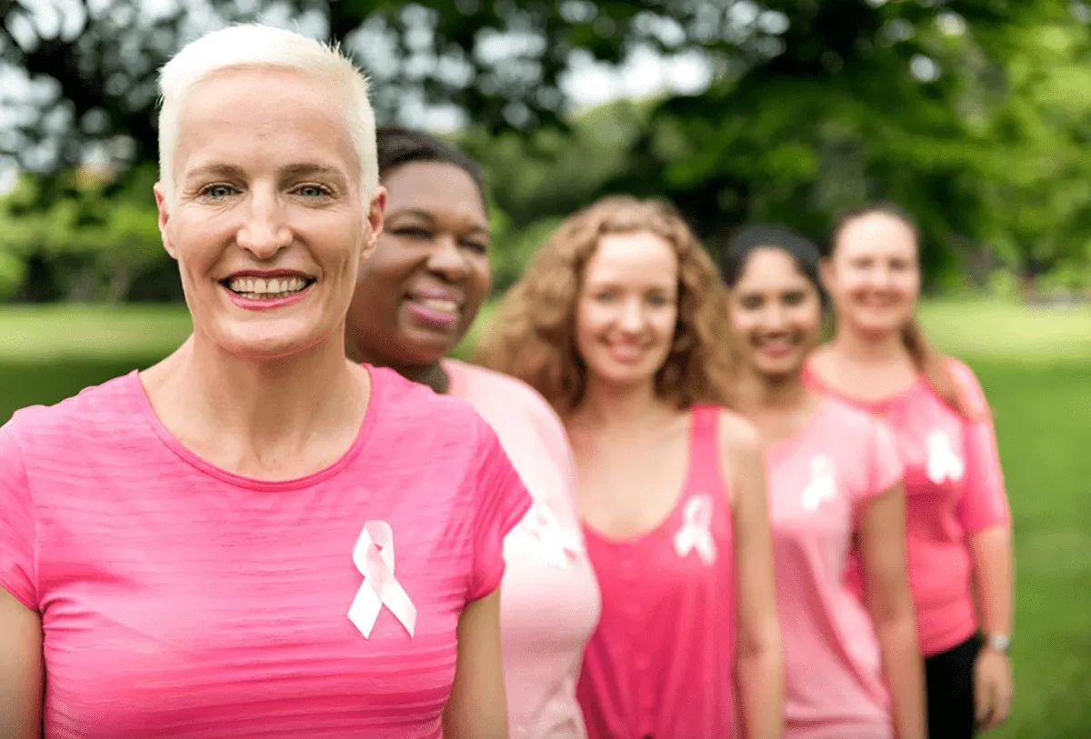 Five women wearing pink tops with breast cancer awareness ribbons stand in a line outdoors at MorningStar Senior Living Hillsboro, smiling, with trees and green grass in the background. The woman in front has short blonde hair.