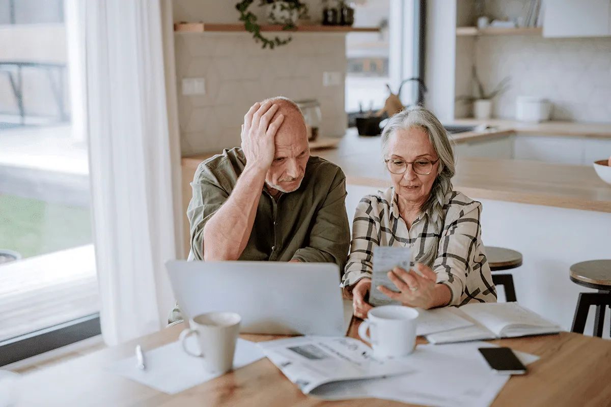 An older man and woman sit at a kitchen table at MorningStar Senior Living Hillsboro, surrounded by a laptop, papers, and coffee cups. The man looks worried, while the woman shows him her phone, both appearing concerned but focused on retirement living plans.