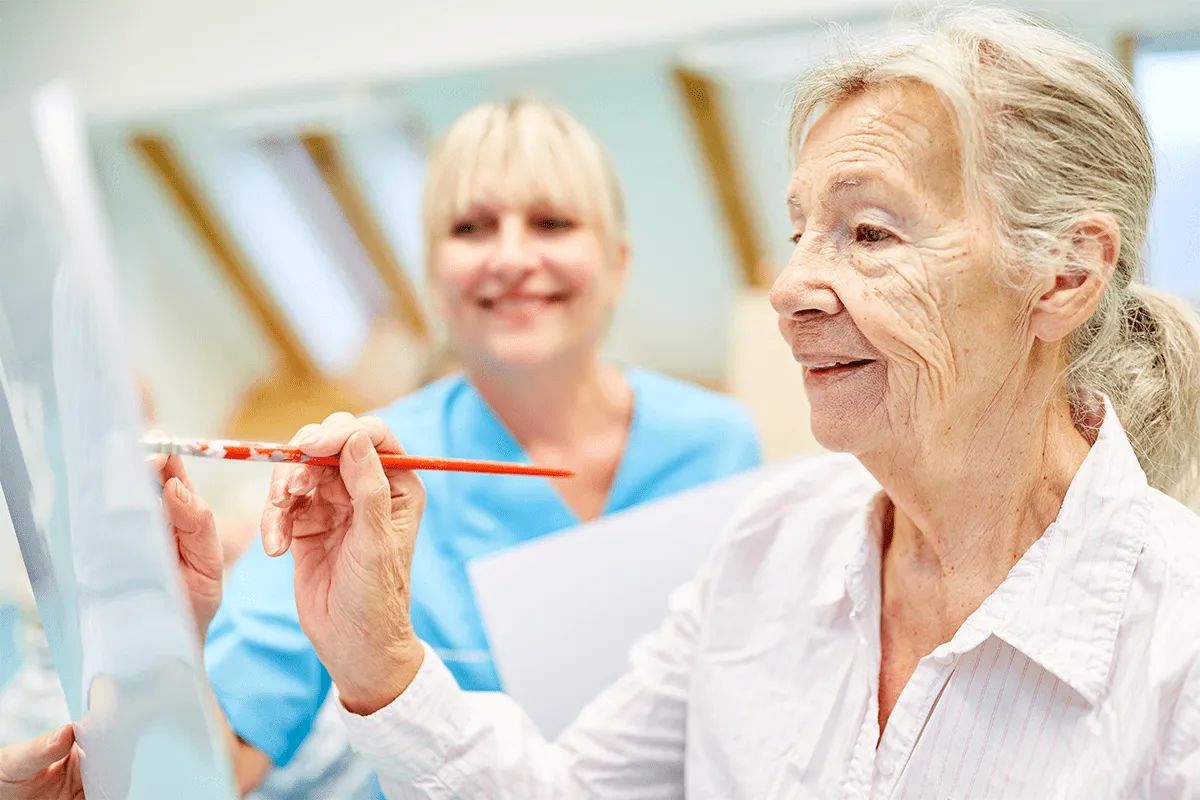 An elderly woman smiles while painting on a canvas with a paintbrush, as a nurse in blue scrubs stands beside her in the bright, welcoming atmosphere of MorningStar Senior Living Hillsboro. Experience the joy of creative retirement living.