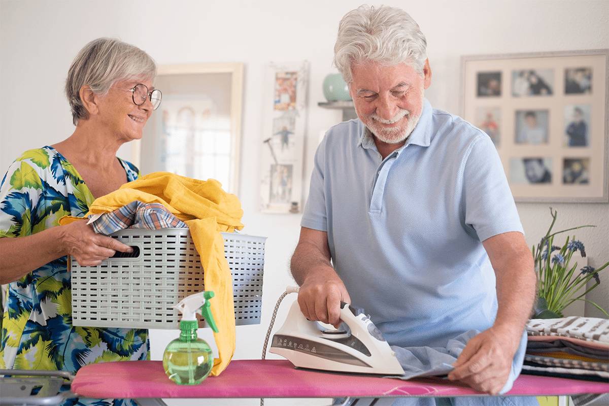 In a bright, homey room at MorningStar Senior Living Hillsboro, an older woman holds a laundry basket and smiles at an older man ironing clothes, showcasing the joys of retirement living together. Folded laundry and a spray bottle rest nearby.