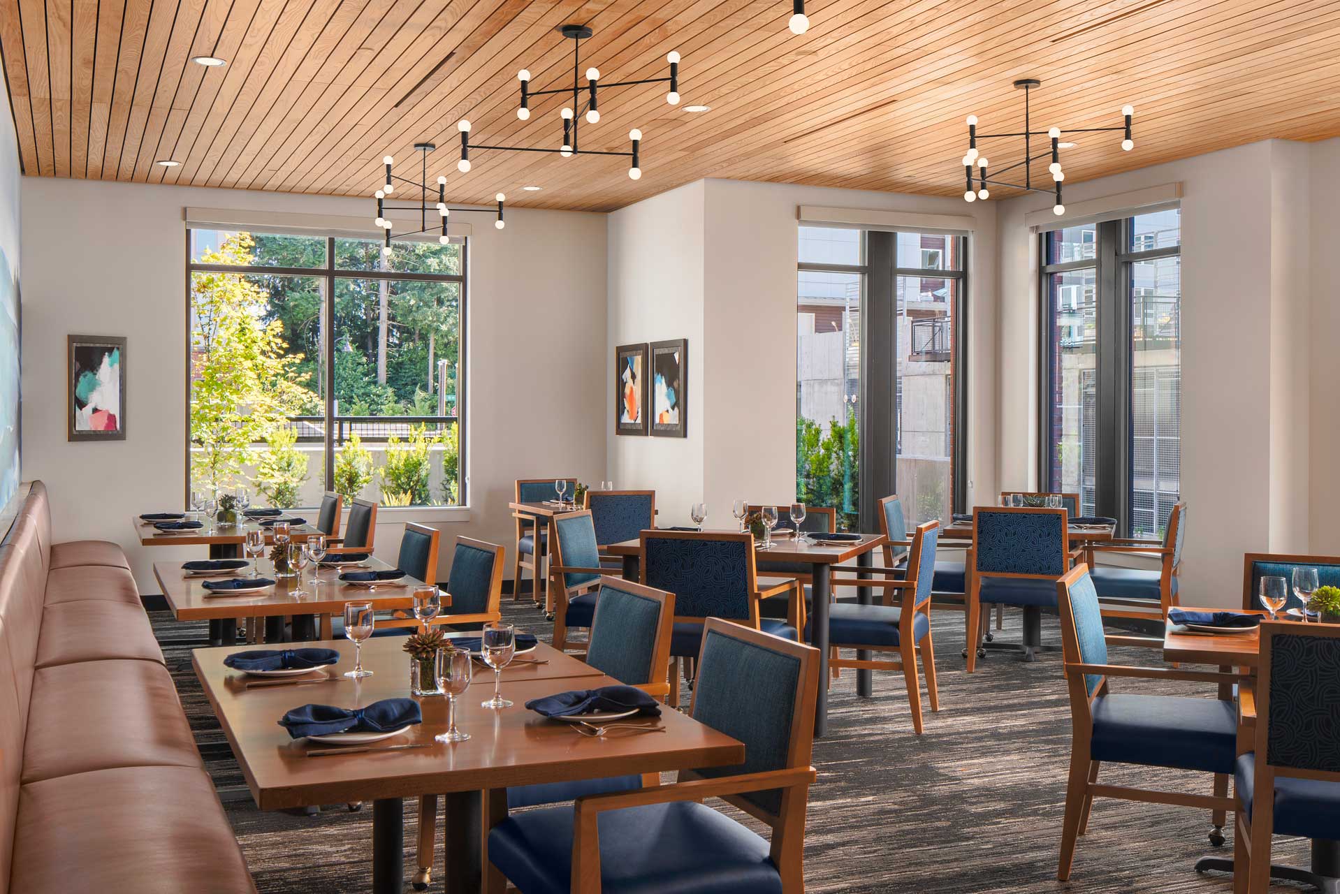 A modern restaurant dining area at MorningStar Senior Living Hillsboro features wooden tables, blue chairs, elegant place settings, and large windows for natural light—an inviting space ideal for enjoying retirement living with style.