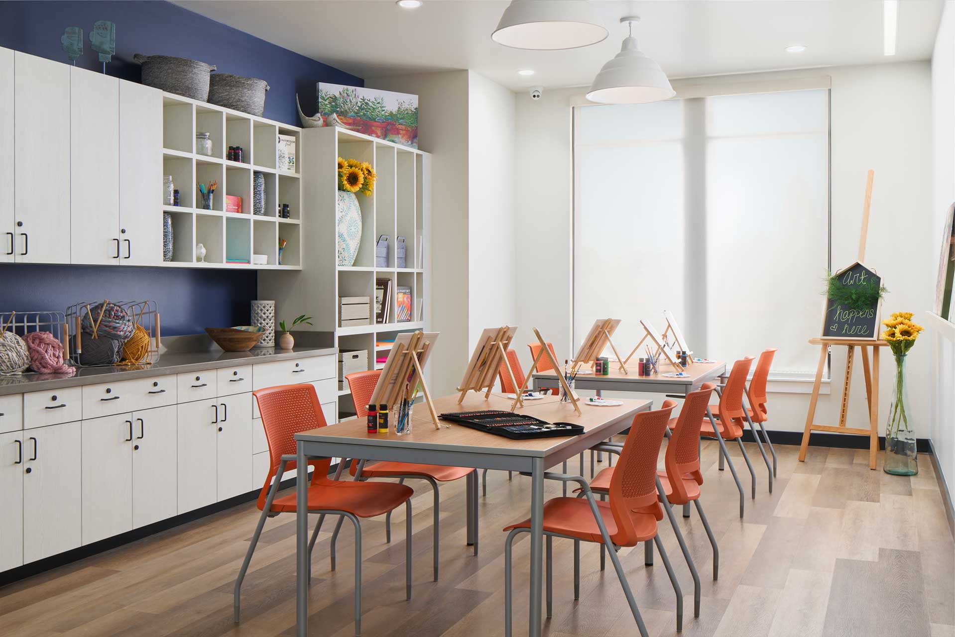 A bright art classroom at MorningStar Senior Living Hillsboro features orange chairs, easels, and painting supplies on tables. White cabinets and shelves with baskets, sunflowers, and art materials line the back wall. Large windows fill the Retirement Living space with light.