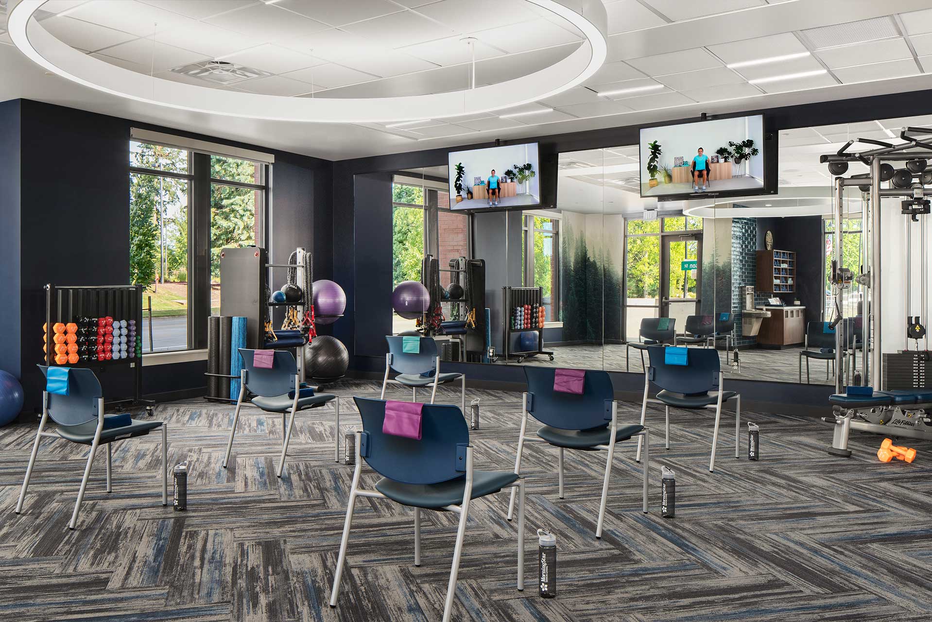 A spacious, modern fitness studio at MorningStar Senior Living Hillsboro features chairs spaced apart facing mirrors, towels on each, and exercise equipment along the walls—perfect for retirement living. Large windows fill the room with natural light.