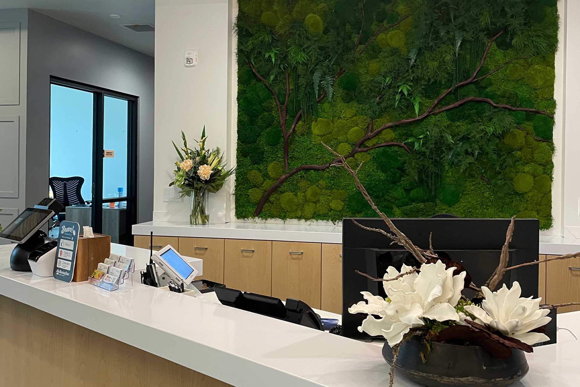 A modern reception desk at MorningStar Senior Living Hillsboro features a white countertop, flower arrangements, and office supplies. A large green plant wall art, light wood cabinets, and natural decor create a welcoming retirement living atmosphere.
