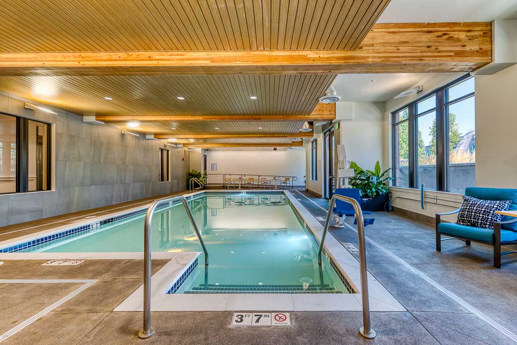 The indoor swimming pool at MorningStar Senior Living Hillsboro features wooden ceiling beams, large windows, and potted plants. Poolside chairs and tables create a relaxing retirement living space, with metal handrails and a 3 ft 7 in depth marker.
