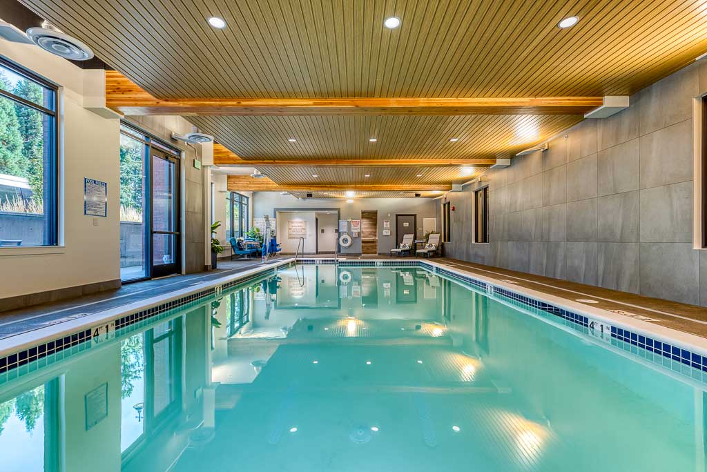 Indoor swimming pool with clear water, surrounded by beige tile flooring and modern gray walls at MorningStar Senior Living Hillsboro. Large windows fill the space with light, creating an inviting retreat for Retirement Living. Lounge chairs await at the far end.