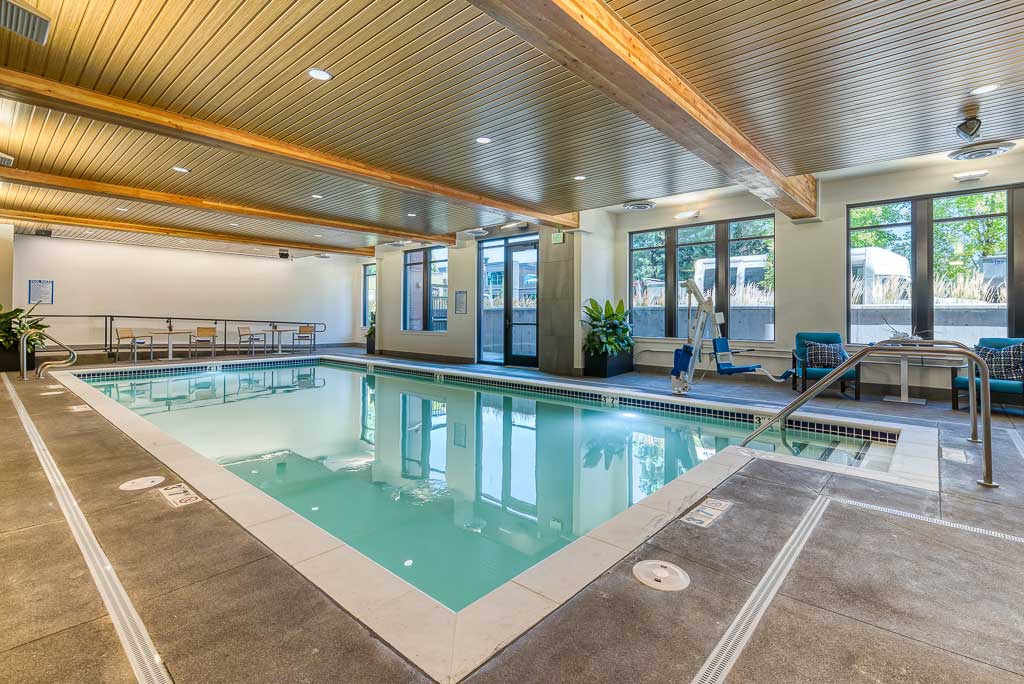 Modern indoor swimming pool at MorningStar Senior Living Hillsboro with beige tile deck, wood beam ceiling, large windows, pool lift, and seating areas. Perfect for relaxation and Retirement Living filled with natural light.