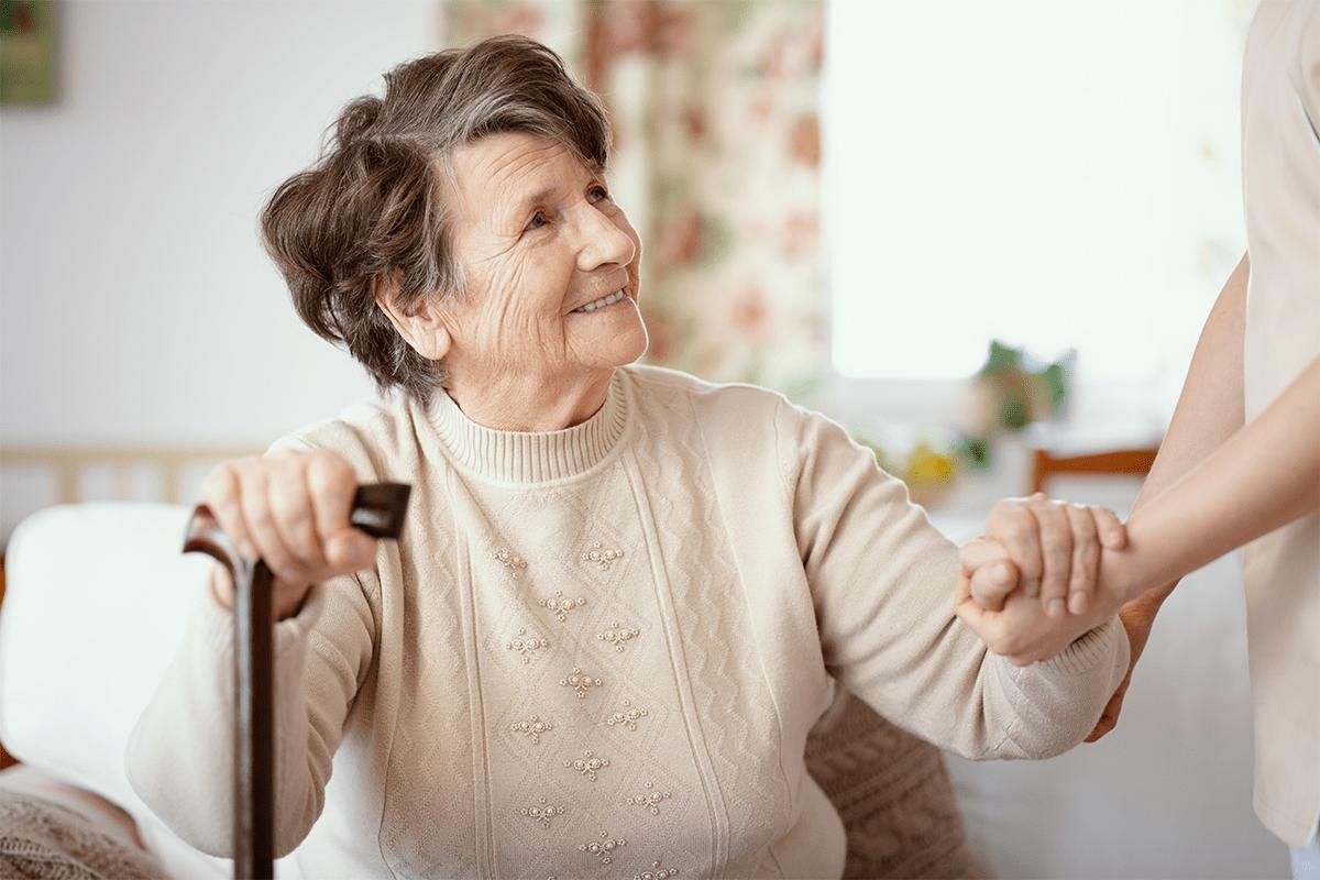 An elderly woman with short gray hair and a cream sweater smiles and holds a cane in one hand while holding the hand of another person, reflecting the supportive community found at MorningStar Senior Living Hillsboro in a cozy, softly lit room.