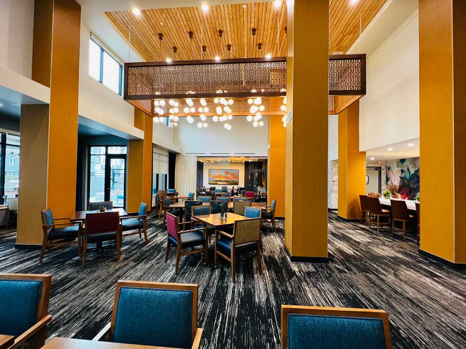 Modern hotel dining area at MorningStar Senior Living Hillsboro, featuring wooden tables, colorful cushioned chairs, tall yellow pillars, patterned lighting fixtures, and large windows welcoming natural light—ideal for vibrant retirement living.