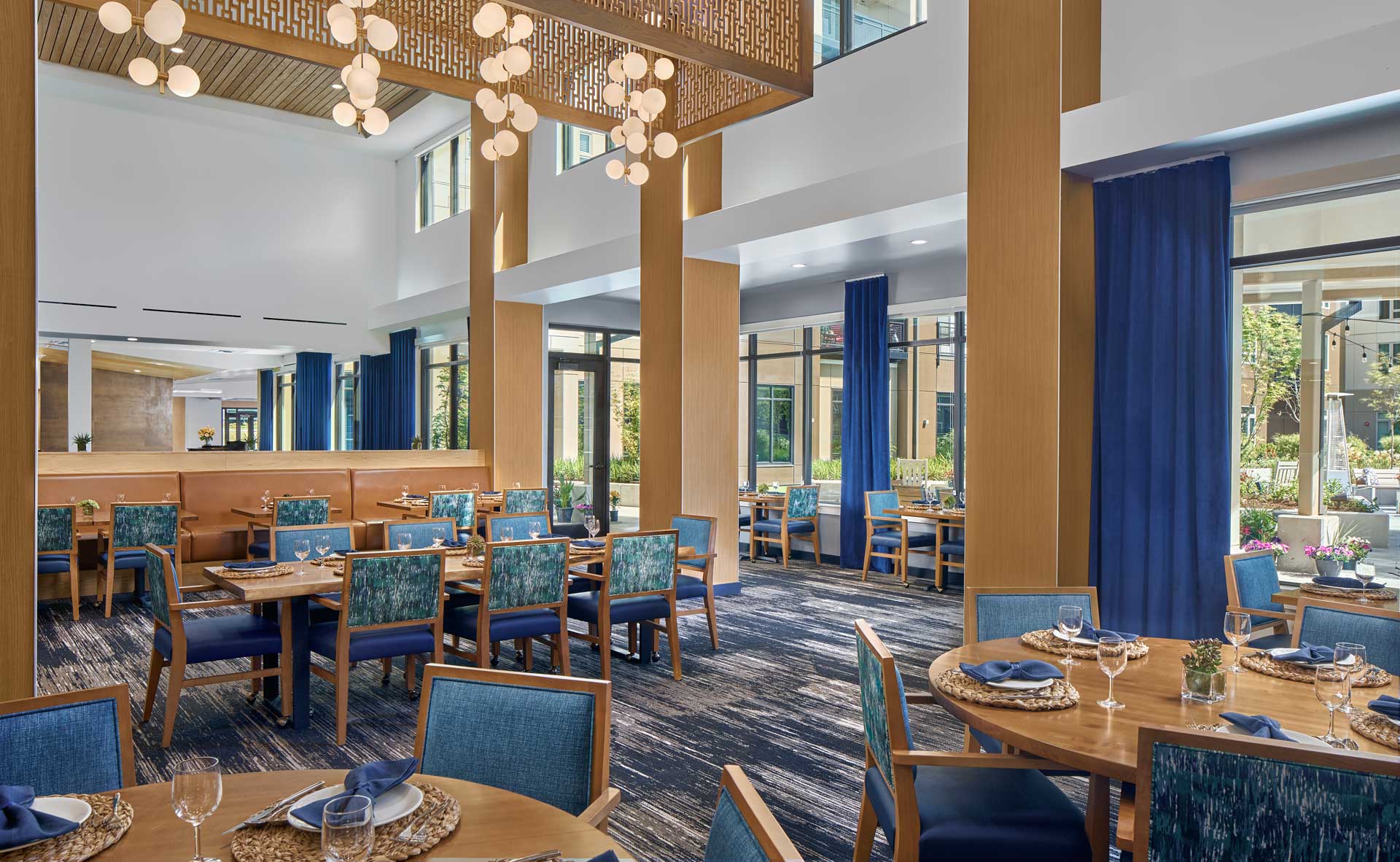 Modern restaurant interior at MorningStar Senior Living Hillsboro features blue and wooden decor, round tables set with plates and napkins, large windows with blue curtains, and contemporary lighting—creating an inviting space for retirement living.