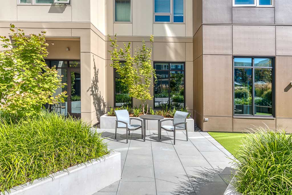 A sunny outdoor patio at MorningStar Senior Living Hillsboro features two modern chairs and a small table, surrounded by green plants and trees, with large windows and beige and brown building walls in the background—a perfect spot for retirement living.