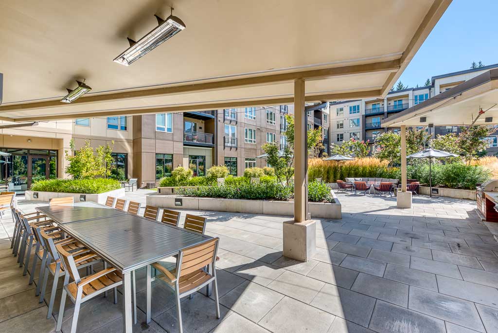 Modern outdoor patio area at MorningStar Senior Living Hillsboro with a long metal dining table and chairs beneath a canopy, surrounded by planters, greenery, and multi-story apartment buildings—ideal for vibrant retirement living.