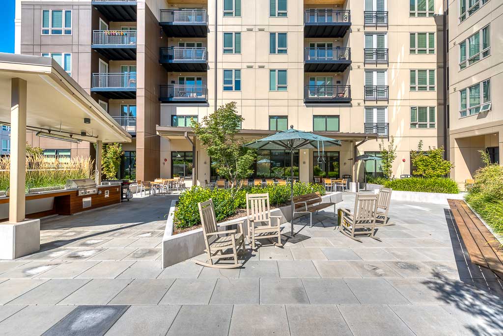 Outdoor courtyard of MorningStar Senior Living Hillsboro features patio seating, rocking chairs, an umbrella, grill area, and lush plants. Large windows and balconies complete this vibrant retirement living space.