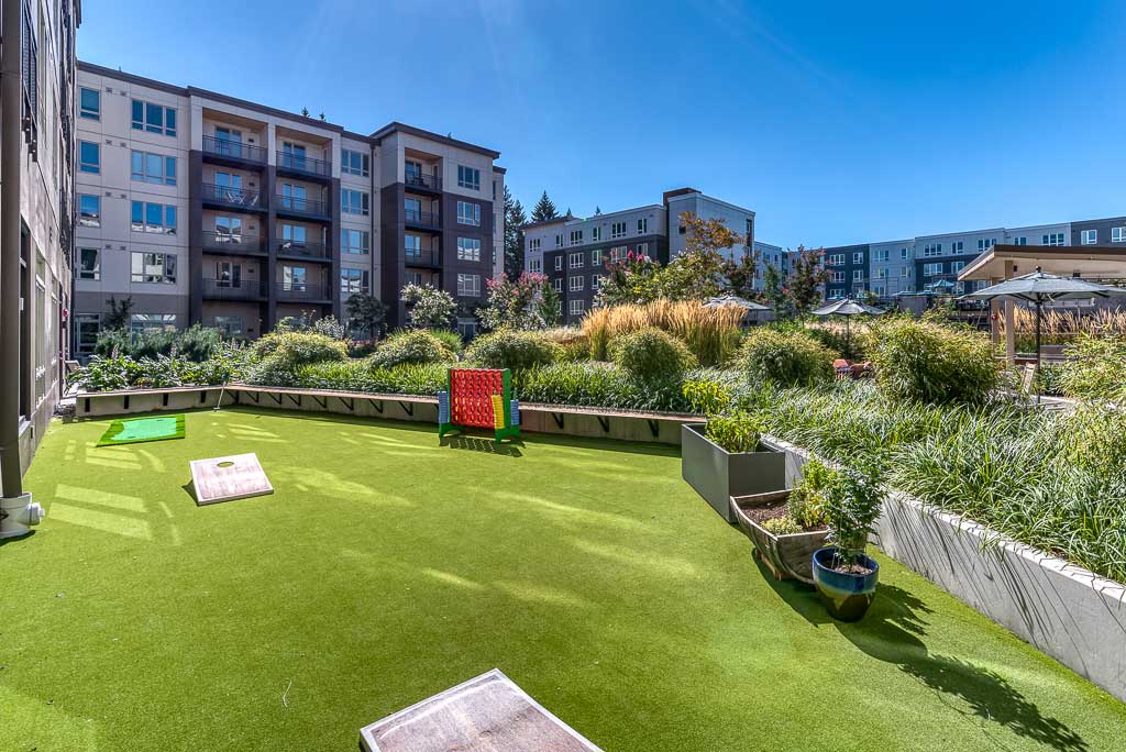 A rooftop courtyard at MorningStar Senior Living Hillsboro features artificial grass, cornhole boards, a large Connect Four game, potted plants, and views of surrounding apartment buildings under a clear blue sky—perfect for retirement living.
