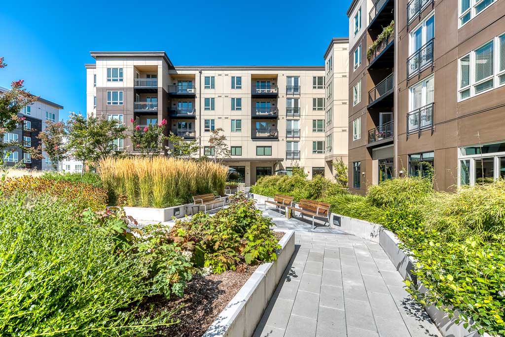 A modern apartment building designed for Retirement Living, with balconies and large windows, surrounds a landscaped courtyard featuring greenery, benches, and a paved walkway under a clear blue sky.