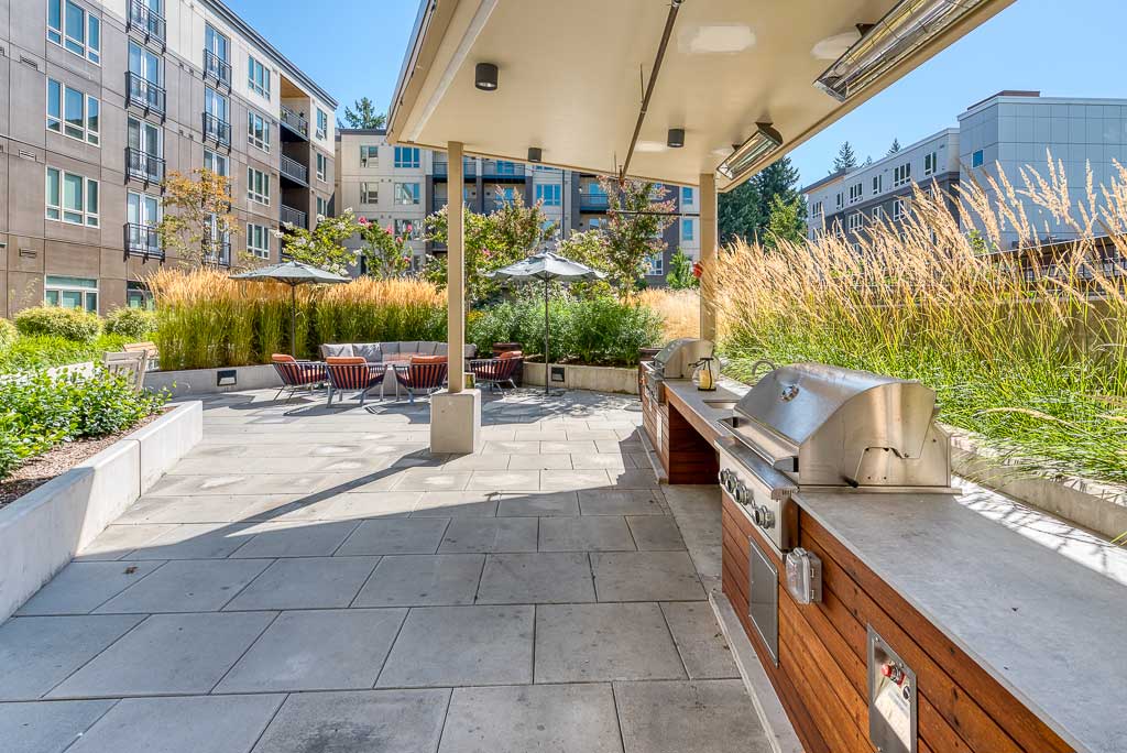 Modern outdoor patio at MorningStar Senior Living Hillsboro features built-in stainless steel grills, wooden cabinets, comfortable seating with umbrellas, and lush grasses—perfect for retirement living on a sunny day amidst apartment buildings.