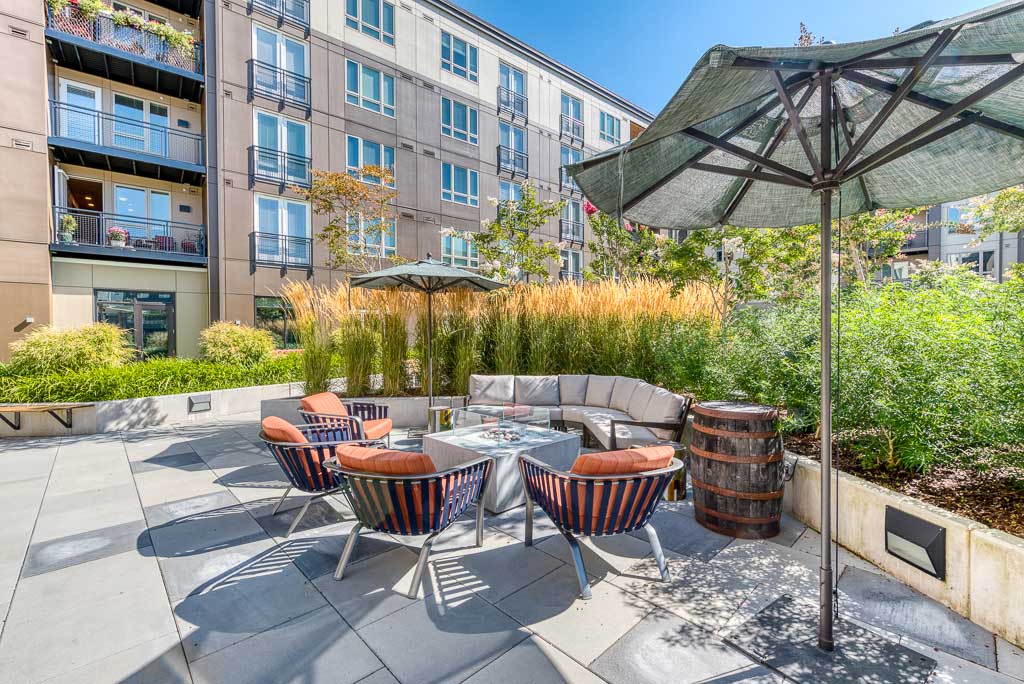 Outdoor patio area with a sectional sofa, round table, striped chairs, umbrellas, and greenery beside a modern apartment—perfect for Retirement Living at MorningStar Senior Living Hillsboro under a clear blue sky.