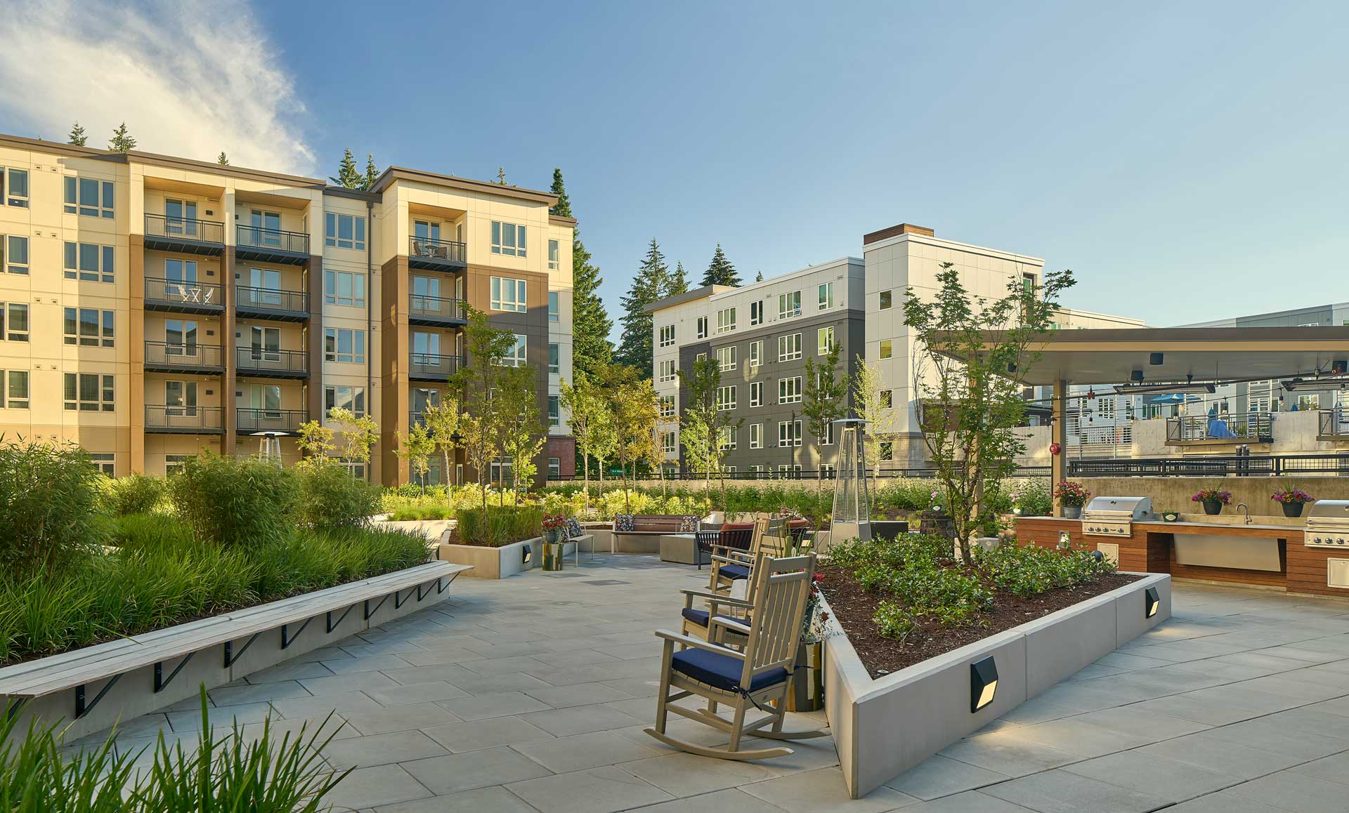 Sunny outdoor patio with benches, rocking chairs, garden planters, and a barbecue grill area at MorningStar Senior Living Hillsboro, surrounded by modern apartment buildings and trees under a clear blue sky—perfect for relaxing retirement living.