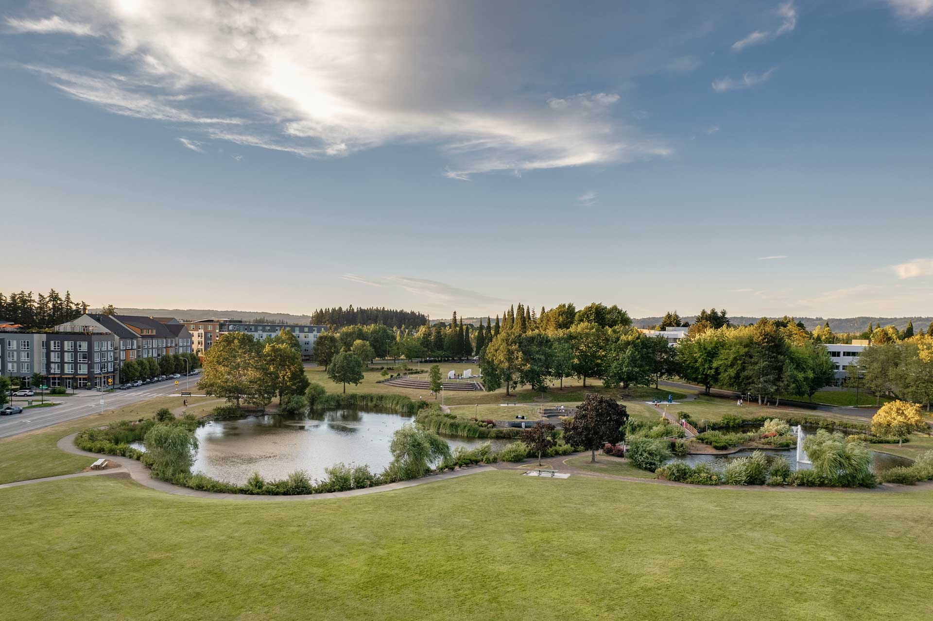 A landscaped park with a pond, fountains, trees, and walking paths welcomes residents of MorningStar Senior Living Hillsboro, surrounded by buildings and roads under a partly cloudy sky—perfect for retirement living.