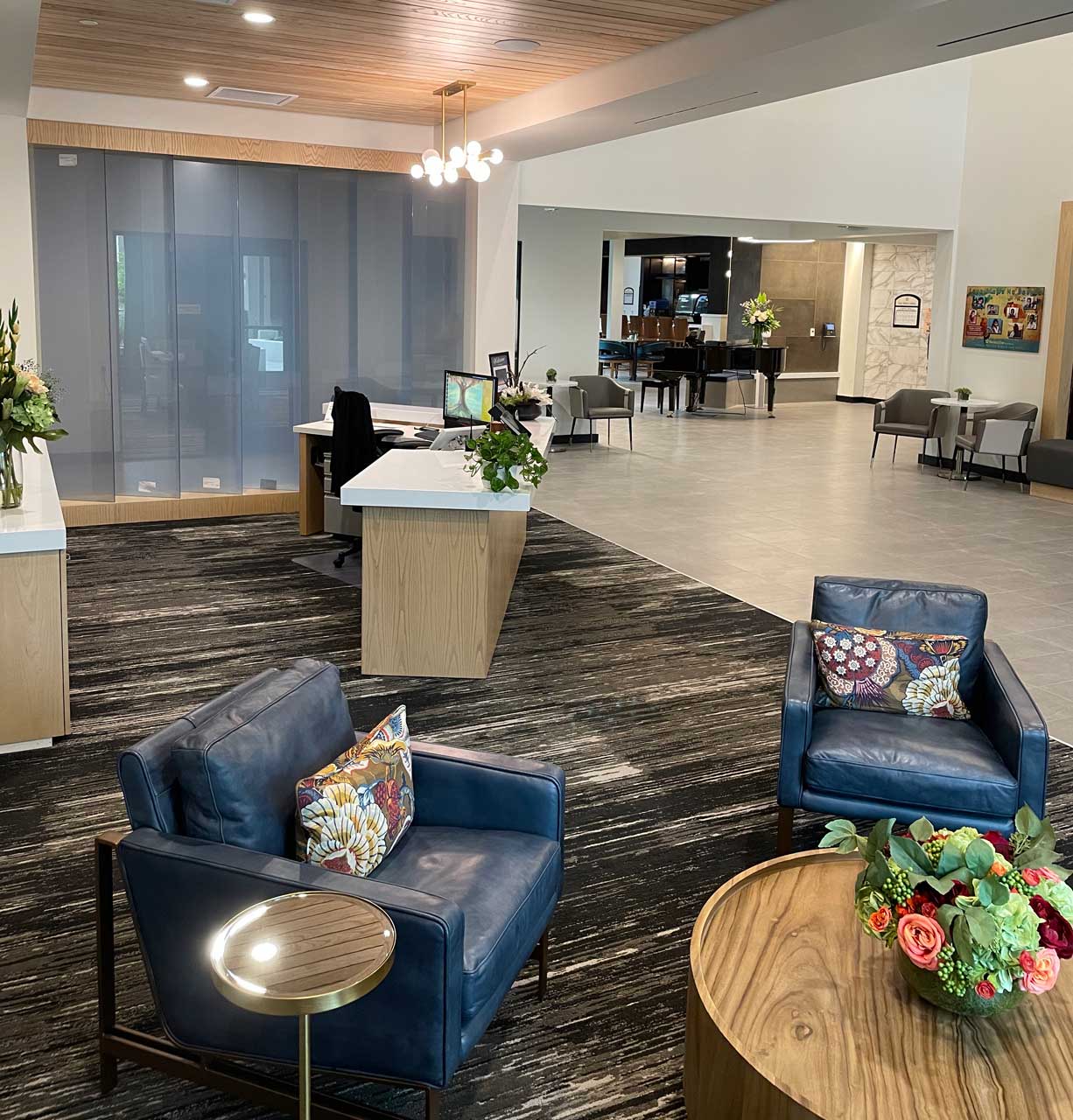 Modern lobby area at MorningStar Senior Living Hillsboro with two blue armchairs, a round wooden table topped with flowers, and a reception desk. Large windows and stylish decor create an inviting space for exceptional retirement living.