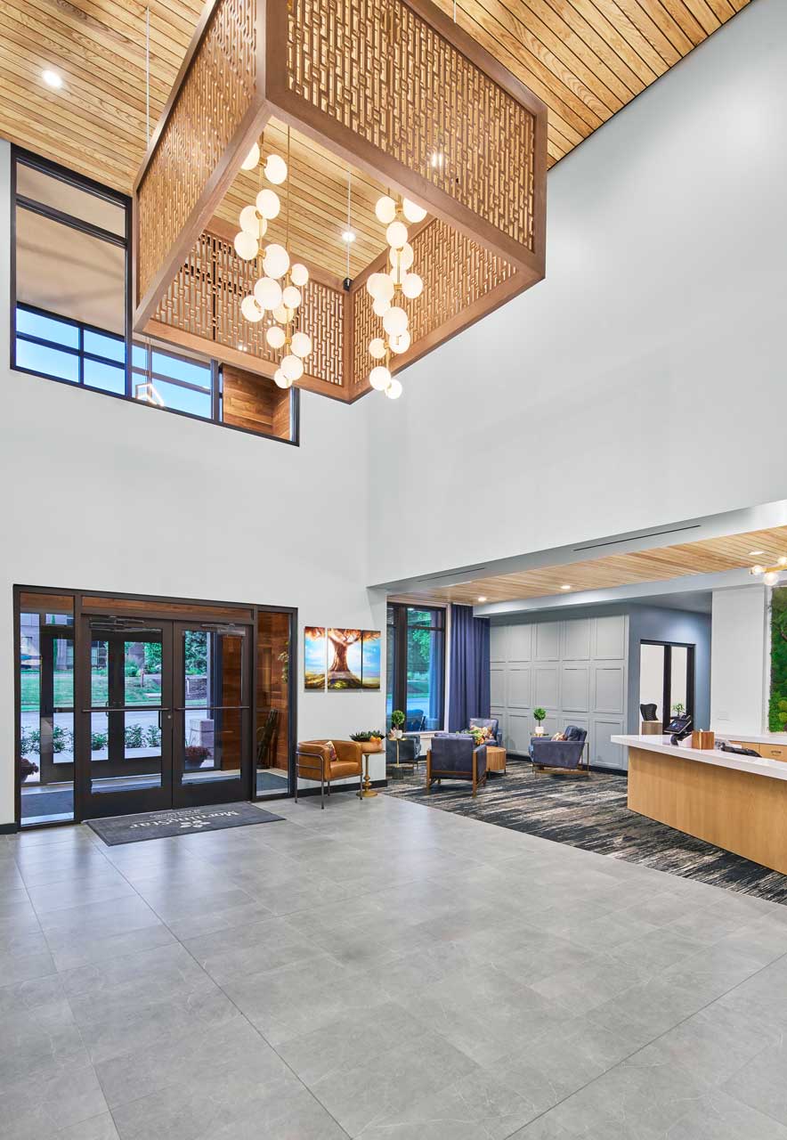 Modern lobby at MorningStar Senior Living Hillsboro with high ceilings, geometric chandelier, large windows, and a reception desk. Seating areas feature chairs, a rug, decor, and plants—offering elegant retirement living near the entrance.