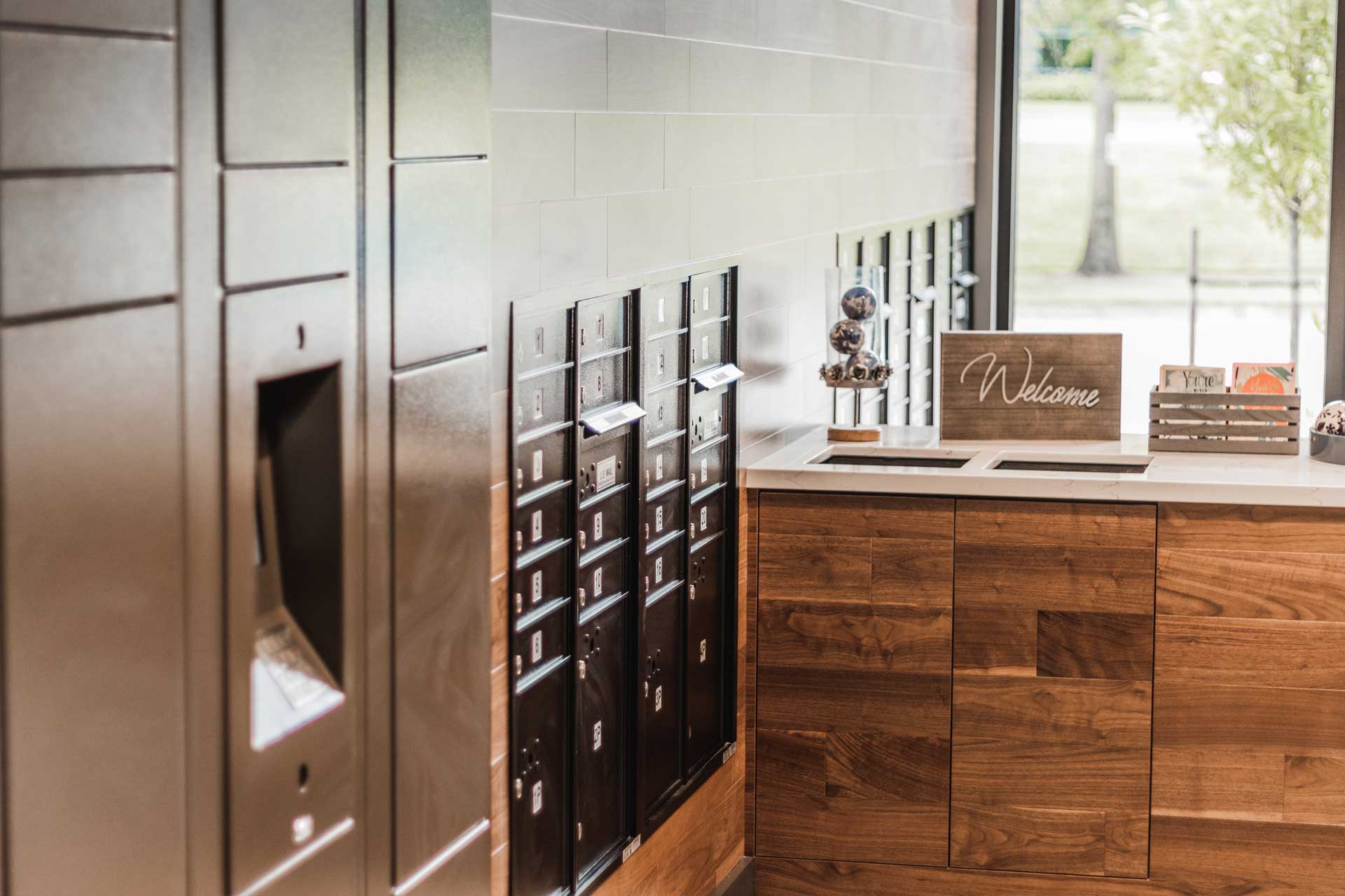 A modern mailroom at MorningStar Senior Living Hillsboro features black mailboxes on the left, wooden cabinets below a white countertop, and a “Welcome” sign displayed near a window with trees visible outside—perfect for comfortable retirement living.