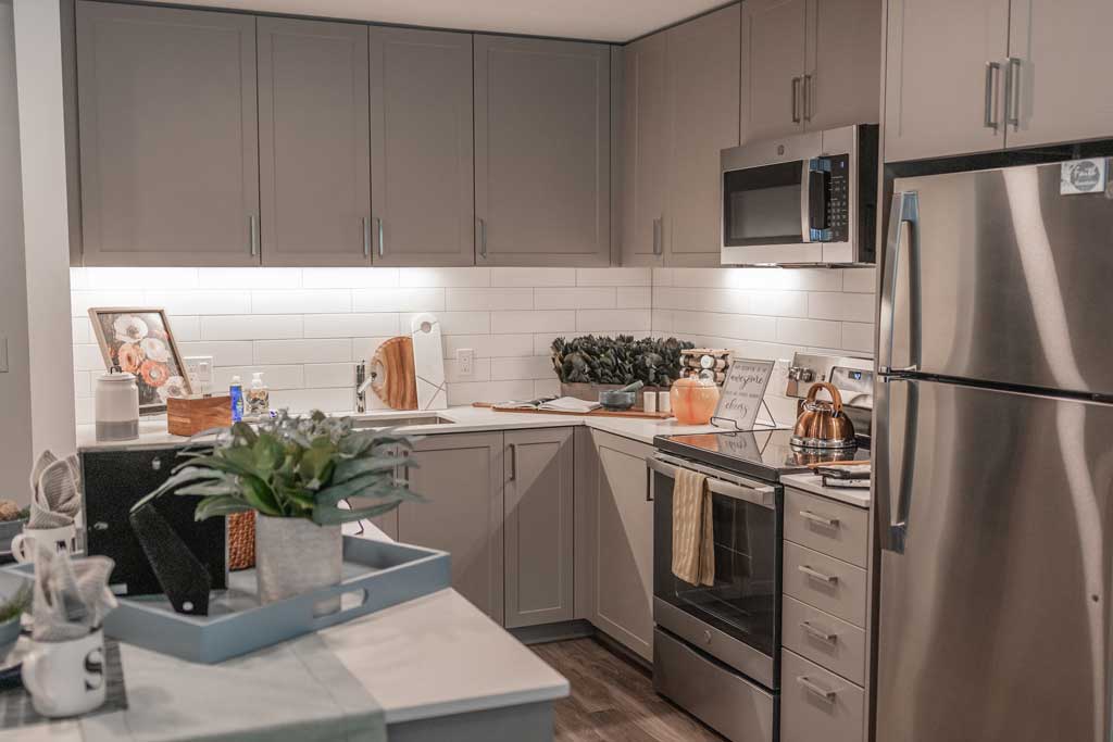Modern kitchen at MorningStar Senior Living Hillsboro with gray cabinets, stainless steel appliances, a kettle on the stove, plants on the counter, and a tray on a white island—perfect for comfortable retirement living. White subway tile and wood floors complete the look.