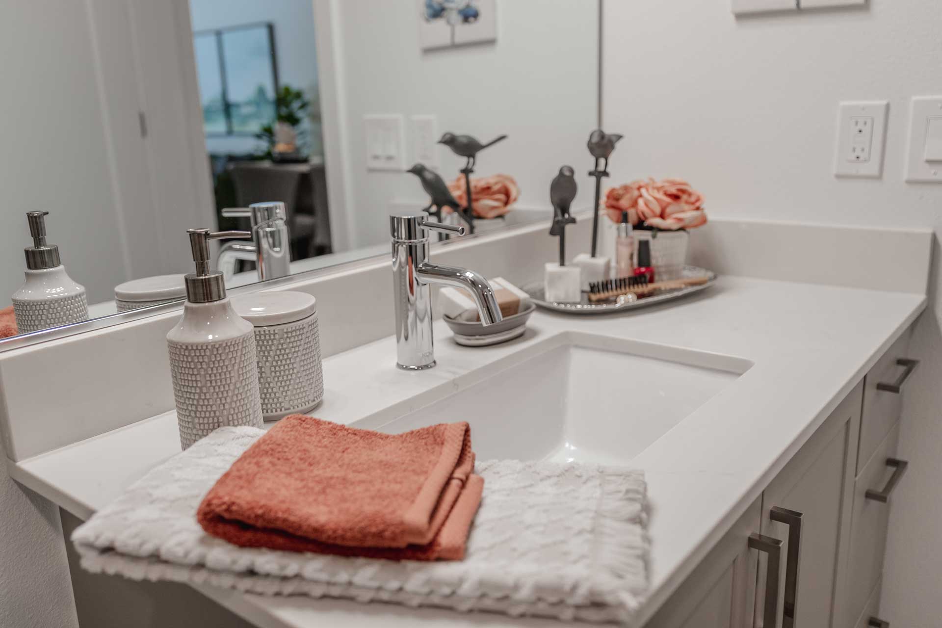 A modern bathroom countertop at MorningStar Senior Living Hillsboro features a white sink, silver faucet, textured containers, folded orange and white towels, decorative flowers, and a tray with toiletries in front of a large mirror.