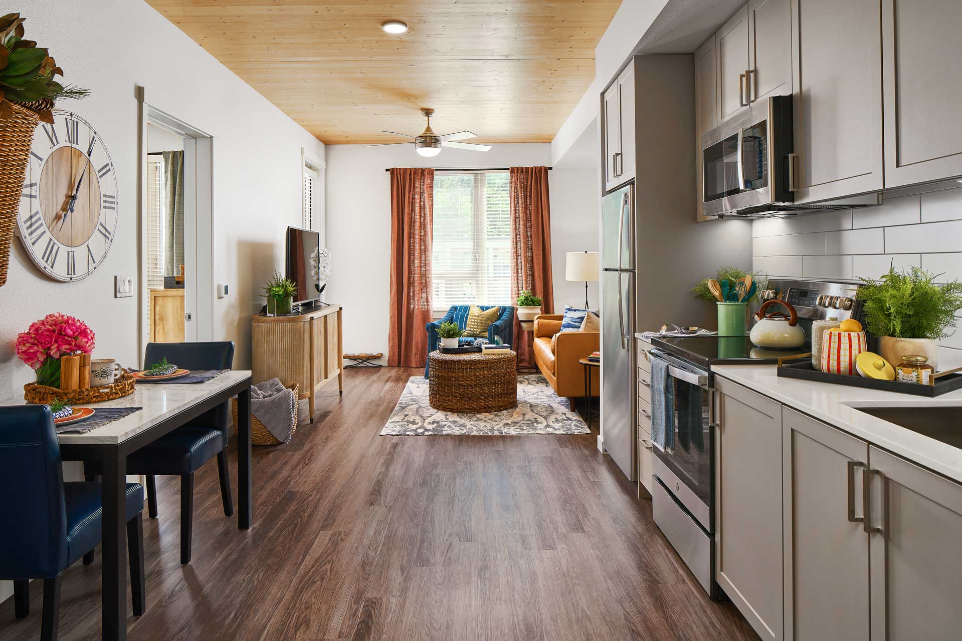Modern apartment interior at MorningStar Senior Living Hillsboro with a kitchen, dining table, and cozy sitting area. Ideal for retirement living, it features grey cabinets, stainless steel appliances, wood floors, and an orange sofa by a large window.