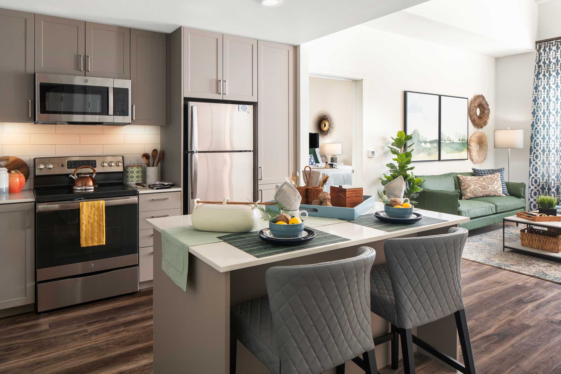 Modern kitchen with gray cabinets, stainless steel appliances, and an island set for two. The kitchen opens to a cozy living room—perfect for Retirement Living at MorningStar Senior Living Hillsboro—with a green sofa, plants, and ample natural light.
