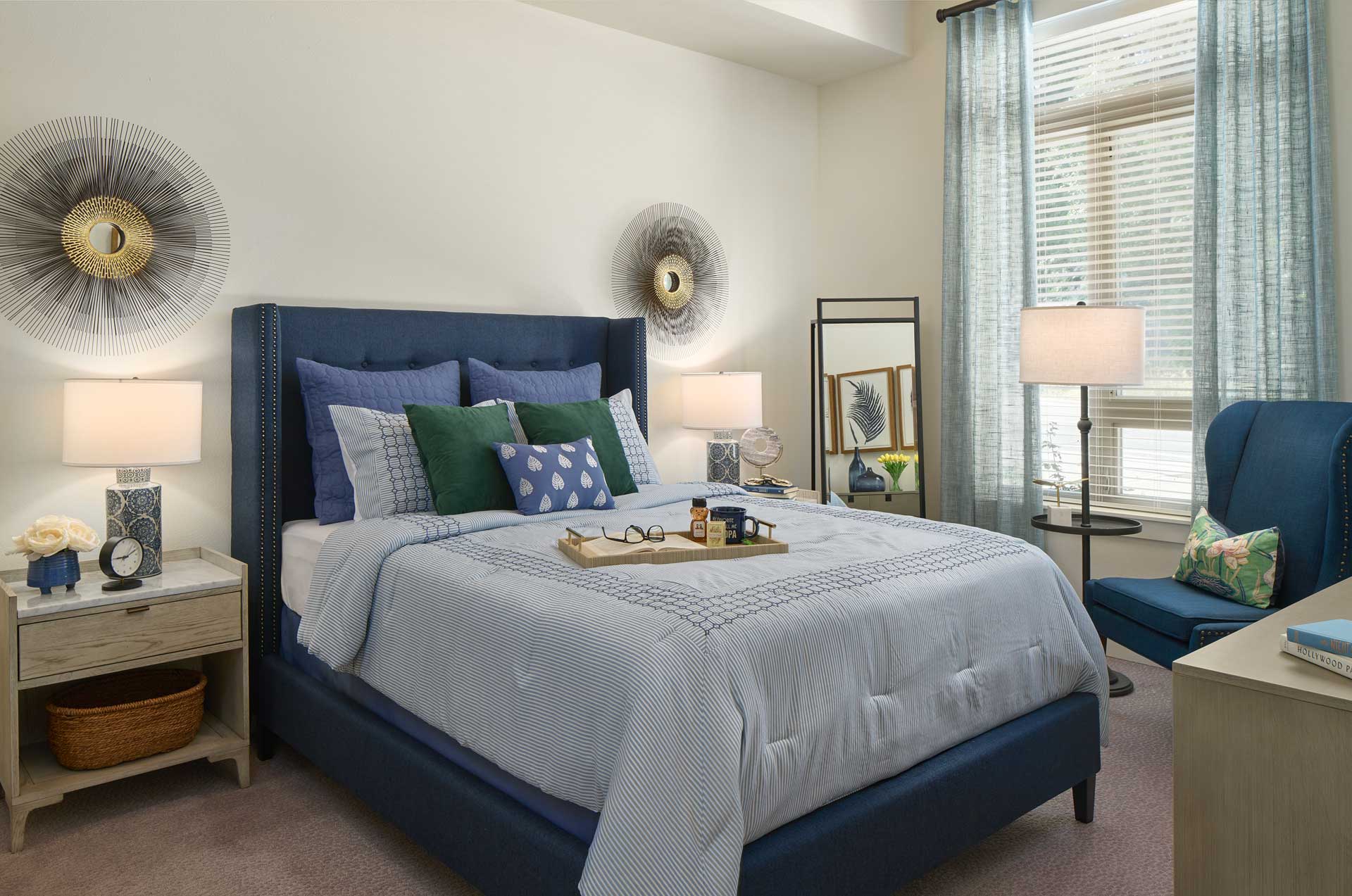 A cozy bedroom at MorningStar Senior Living Hillsboro features a blue upholstered bed, light bedding, and accent pillows. Two nightstands with lamps, wall art above the bed, and sheer curtains create an inviting retreat for retirement living.