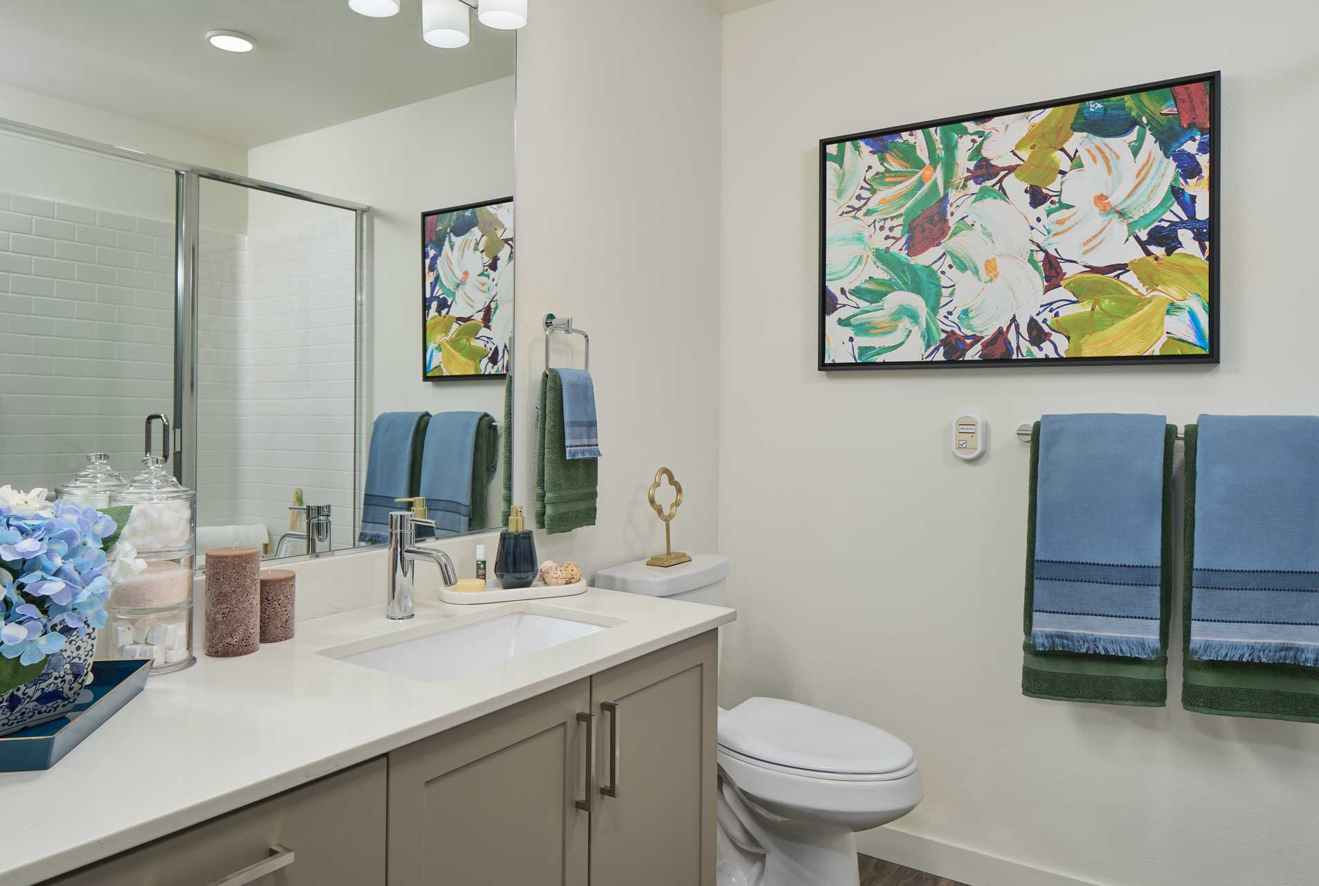 Modern bathroom at MorningStar Senior Living Hillsboro features a white countertop, beige cabinets, two blue towels, a glass shower, floral wall art, and decorative touches—perfect for comfortable retirement living.