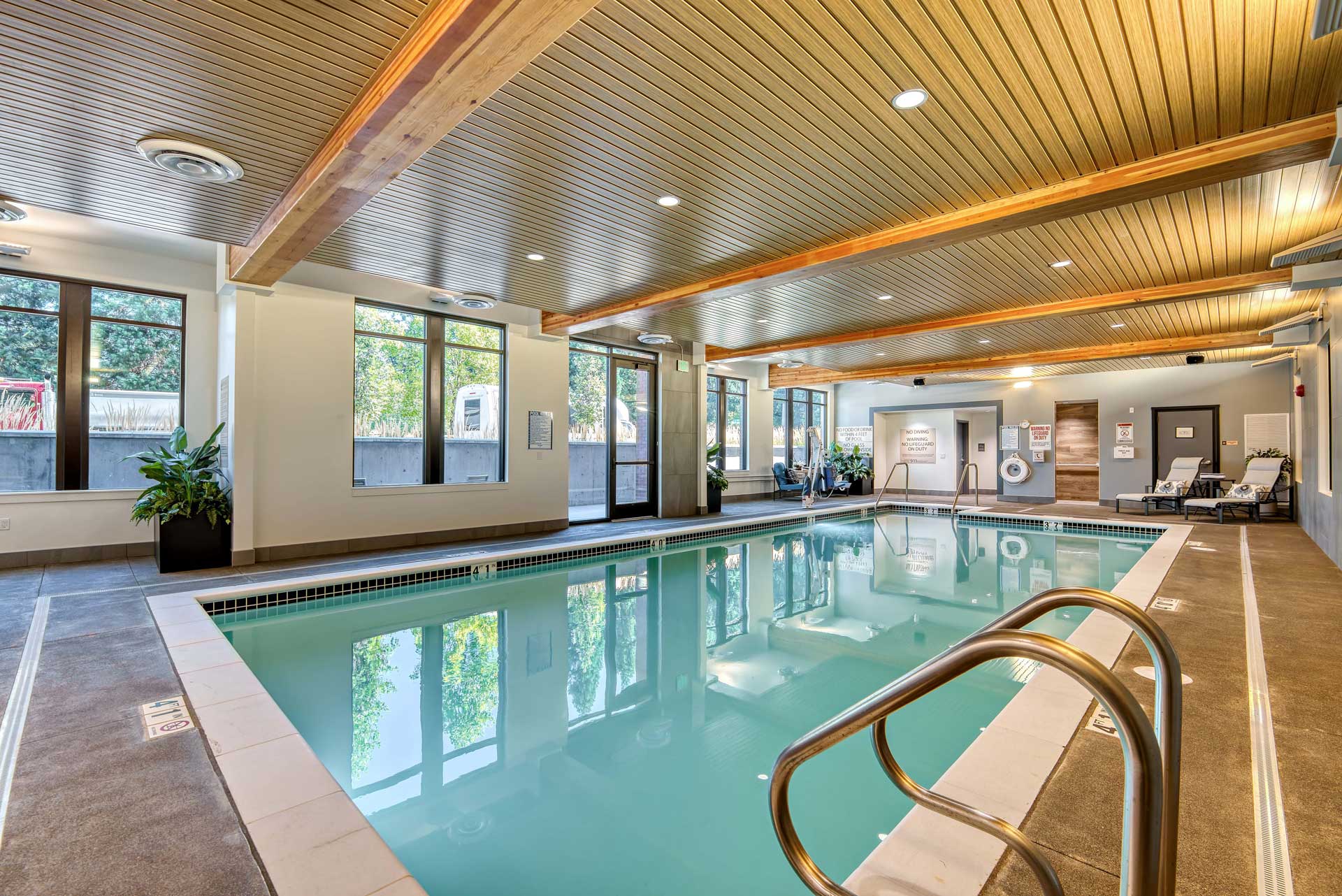Experience Retirement Living at its finest in this modern indoor swimming pool at MorningStar Senior Living Hillsboro, featuring clear water, stainless steel handrails, lounge chairs, lush plants, and sunlit wood-paneled ceilings with exposed beams.