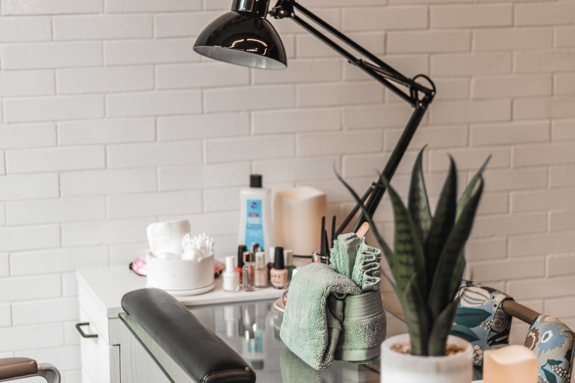 A manicure station with a desk lamp, rolled green towels, nail polish bottles, lotion, cotton pads, and a potted plant, set against a white brick wall—perfect for relaxing moments at MorningStar Senior Living Hillsboro.
