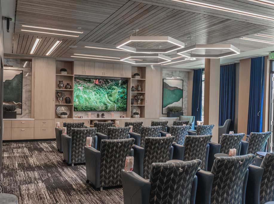 A modern, well-lit room at MorningStar Senior Living Hillsboro features rows of patterned blue armchairs facing a large flat-screen TV. Shelves and artwork decorate the walls, with geometric lights hanging from a wood-paneled ceiling.