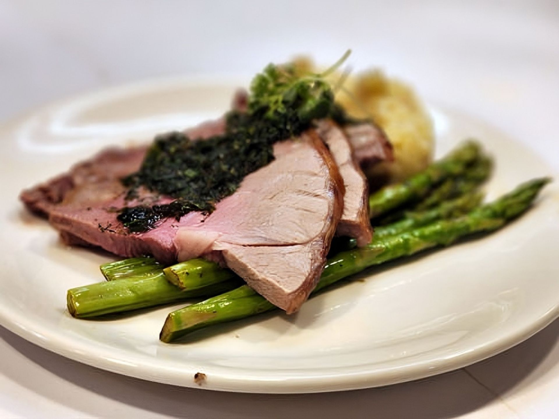Sliced roast meat topped with green herb sauce, served over grilled asparagus spears and mashed potatoes—a delicious example of fine dining at MorningStar Senior Living Hillsboro, setting the standard for exceptional retirement living.