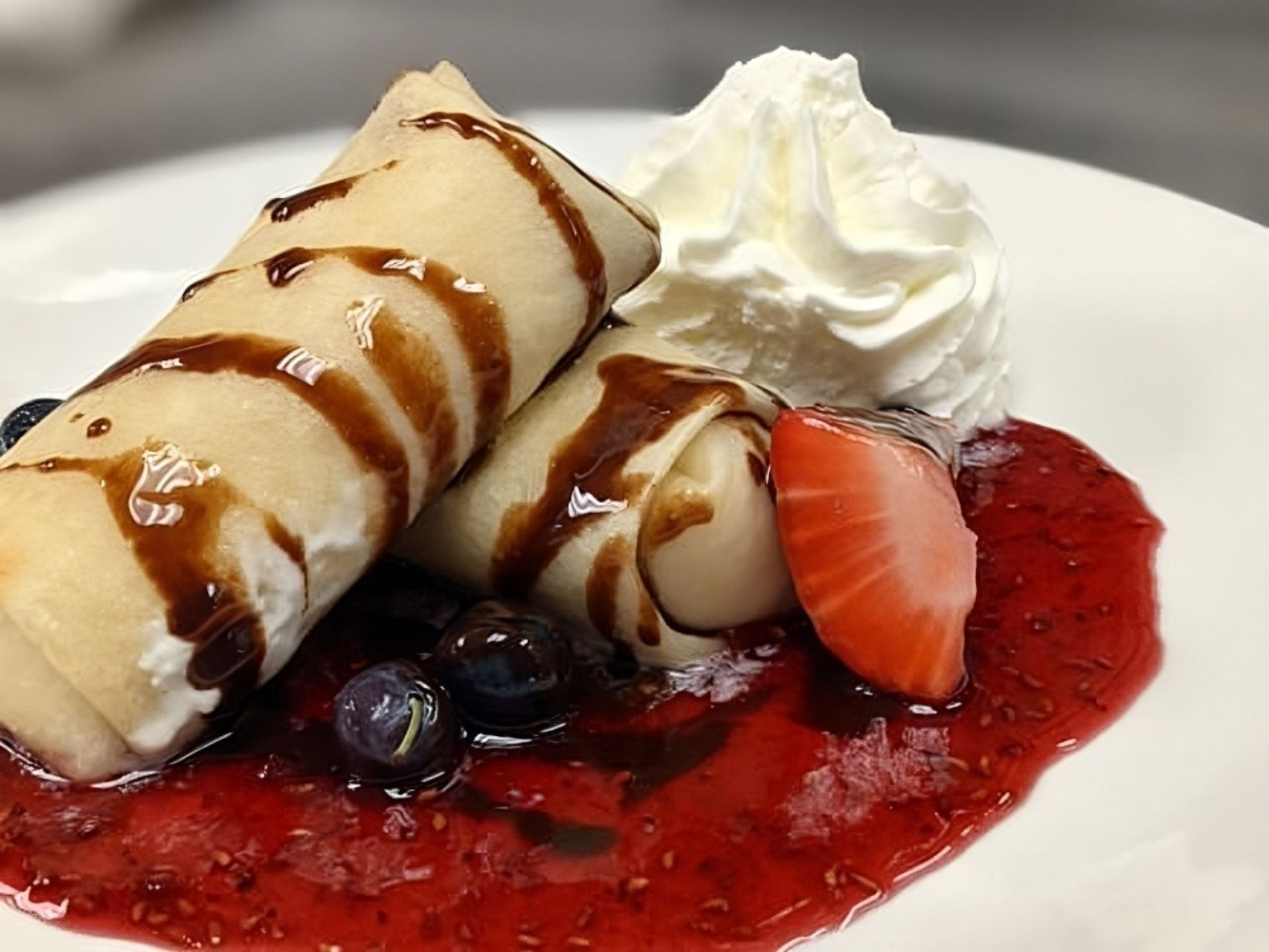 Two rolled crepes drizzled with chocolate sauce, served on a plate with whipped cream, fresh blueberries, a strawberry slice, and red berry sauce—an inviting treat at MorningStar Senior Living Hillsboro for exceptional retirement living.