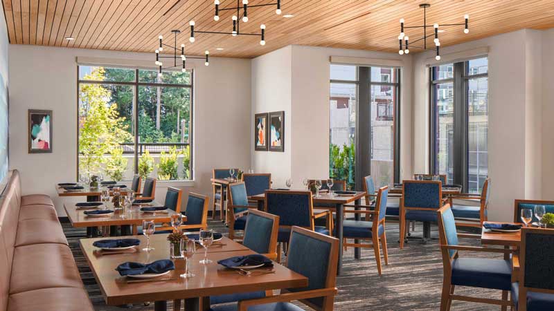 Modern restaurant dining area at MorningStar Senior Living Hillsboro featuring wooden tables, blue cushioned chairs, large windows for natural light, contemporary lighting fixtures, and framed art—perfect for elegant Retirement Living.