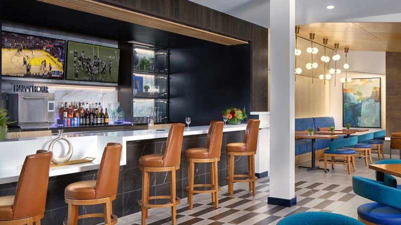 A modern hotel bar at MorningStar Senior Living Hillsboro features tan leather barstools, a white counter, liquor bottles, and TVs showing sports. Blue booth seating and round tables enhance the dining area’s contemporary retirement living atmosphere.