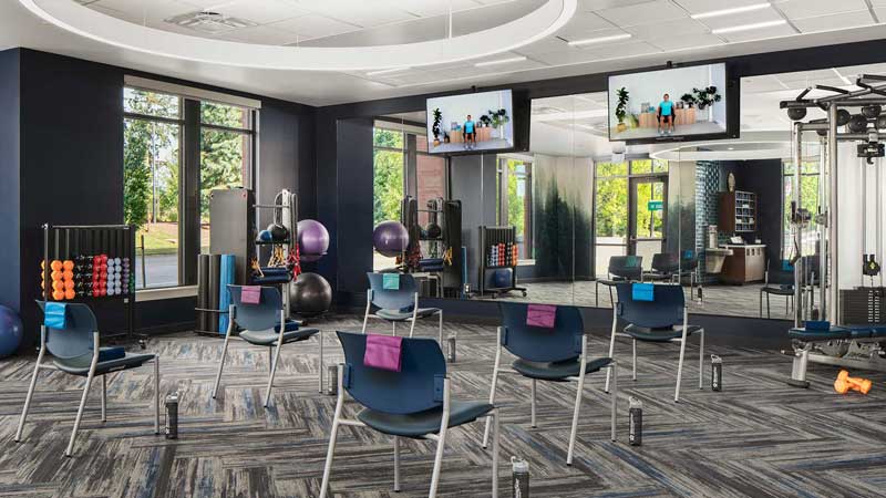 A modern fitness studio at MorningStar Senior Living Hillsboro features chairs spaced apart, each with a towel and water bottle, exercise balls, dumbbells, large windows with views, and TV screens displaying workout videos—perfect for active retirement living.