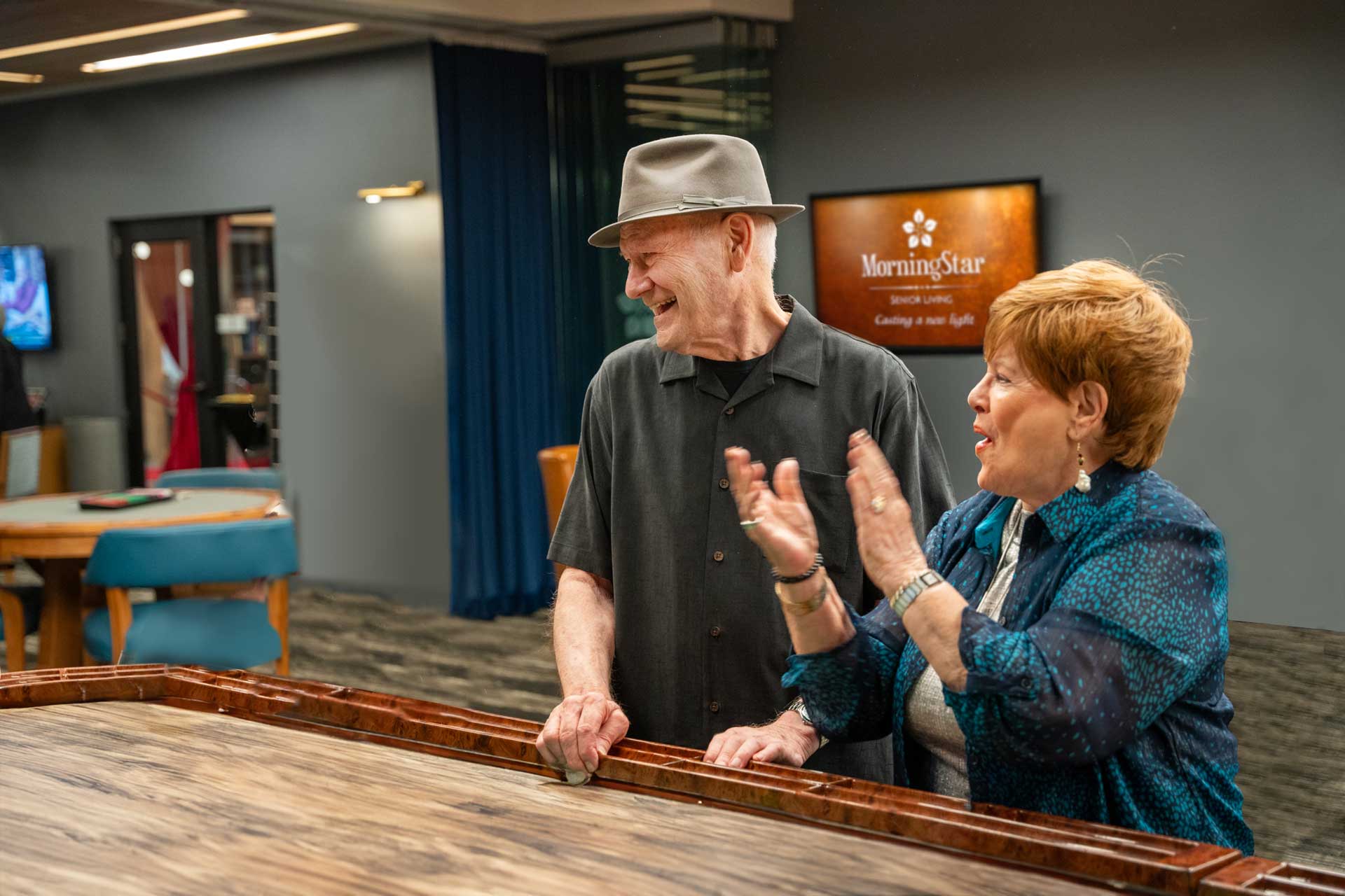 An older man and woman stand at a game table, smiling and laughing together. The woman is clapping her hands. Behind them, a sign reads “MorningStar Senior Living Hillsboro” in a warmly lit retirement living community.