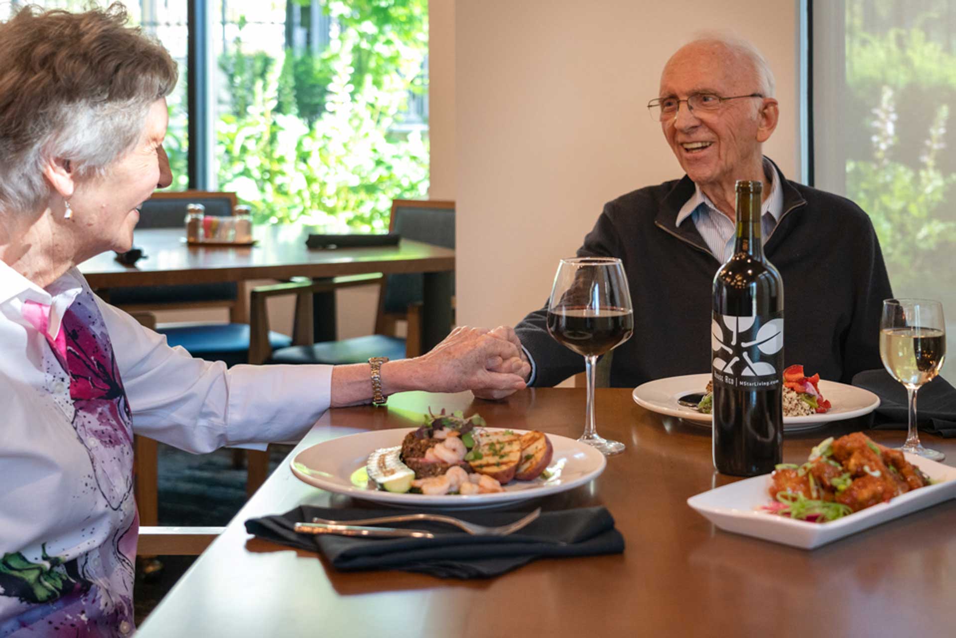 An elderly couple sits at a restaurant table at MorningStar Senior Living Hillsboro, holding hands and smiling. Plates of food, a bottle of wine, and glasses are before them as sunlight streams through the large window behind.